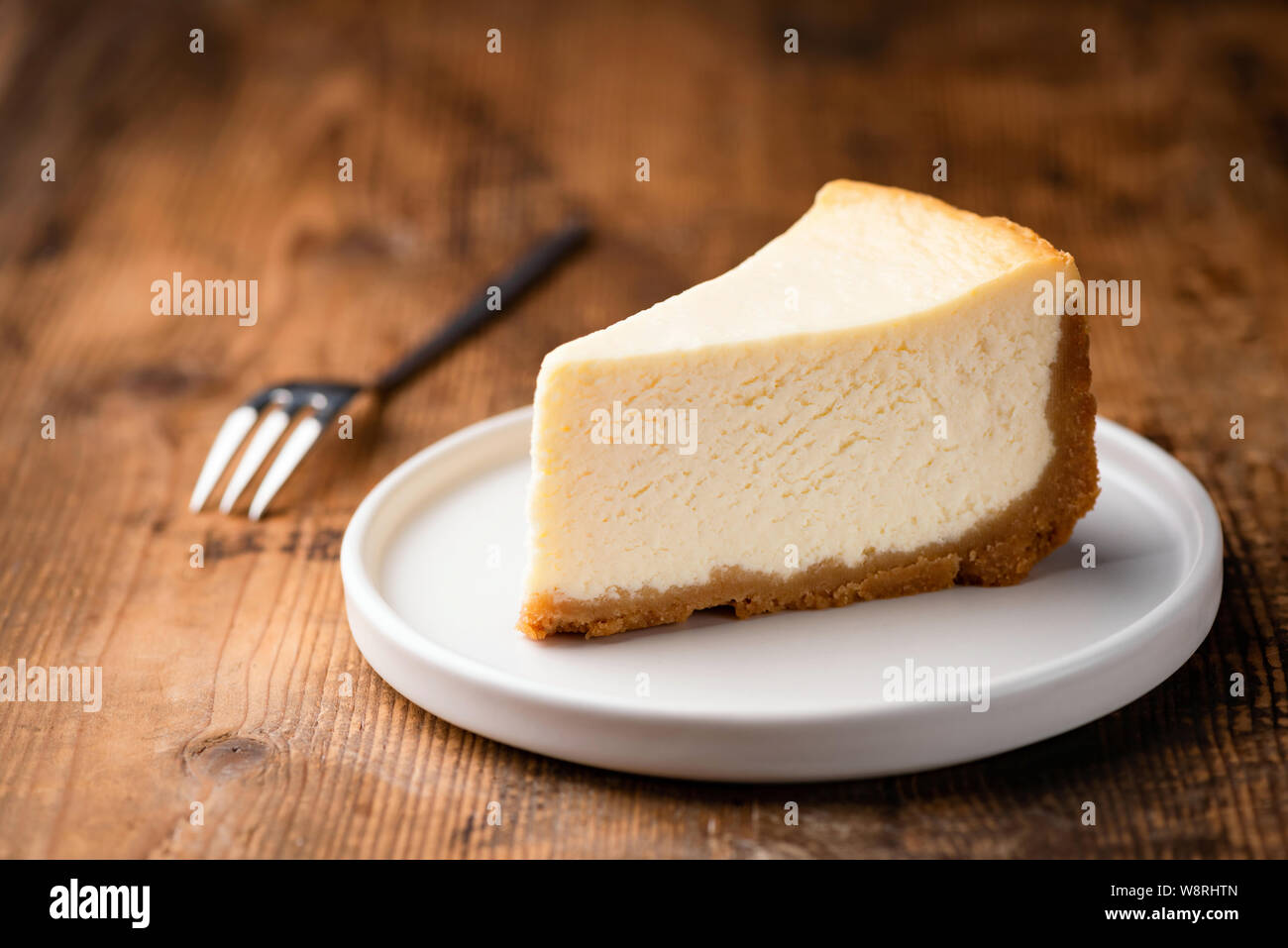 Tarta de queso estilo Nueva York slice, la clásica tarta de queso sobre fondo de madera. Rebanada de pastel sabroso sobre placa blanca servido con horquilla de postre Foto de stock