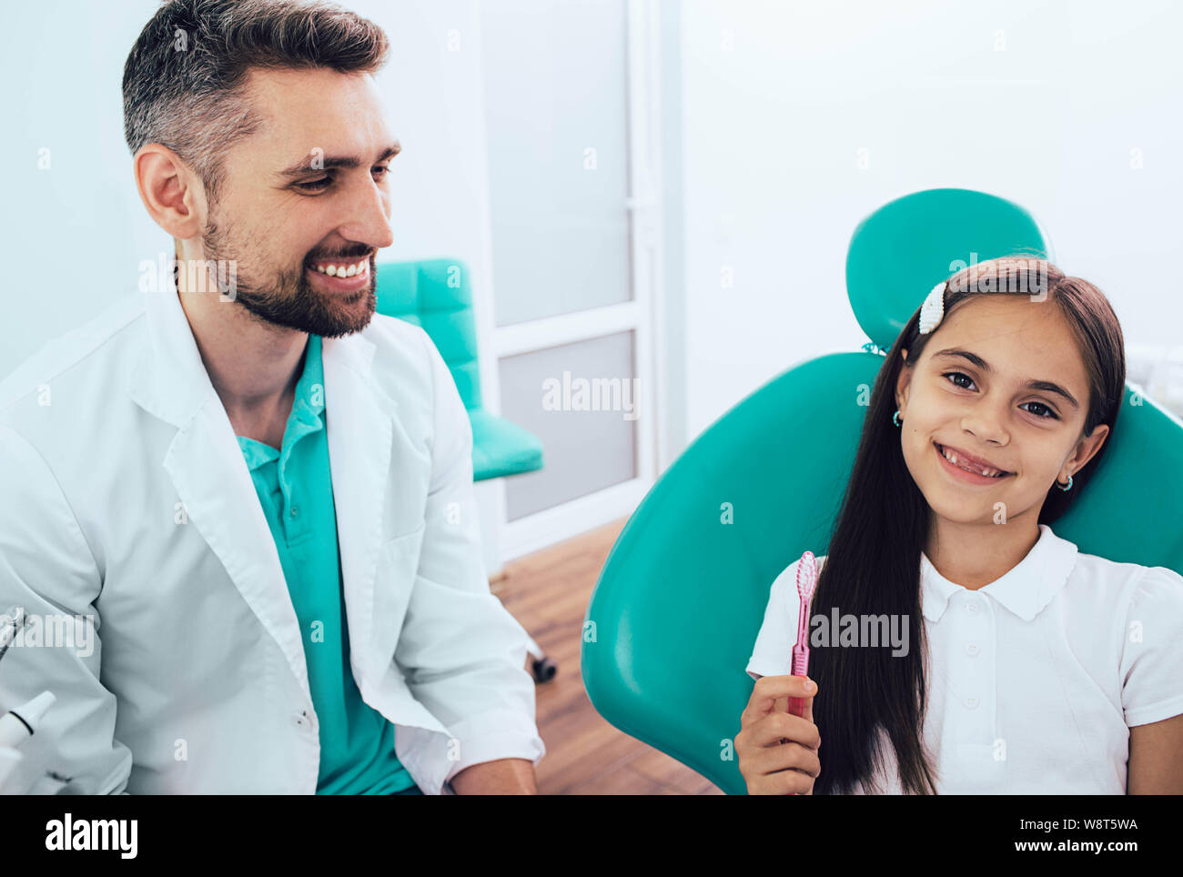 Pequeña niña de raza mixta en el sillón dental sentado cerca de su dentista. Ella sujetando el cepillo dental y sonriente. La forma correcta de cepillado de dientes Foto de stock