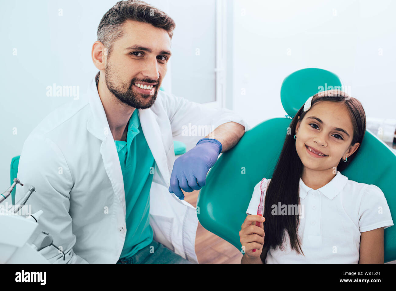 Pequeña niña de raza mixta en el sillón dental sentado cerca de su dentista. Ella sujetando el cepillo dental y sonriente. La forma correcta de cepillado de dientes Foto de stock