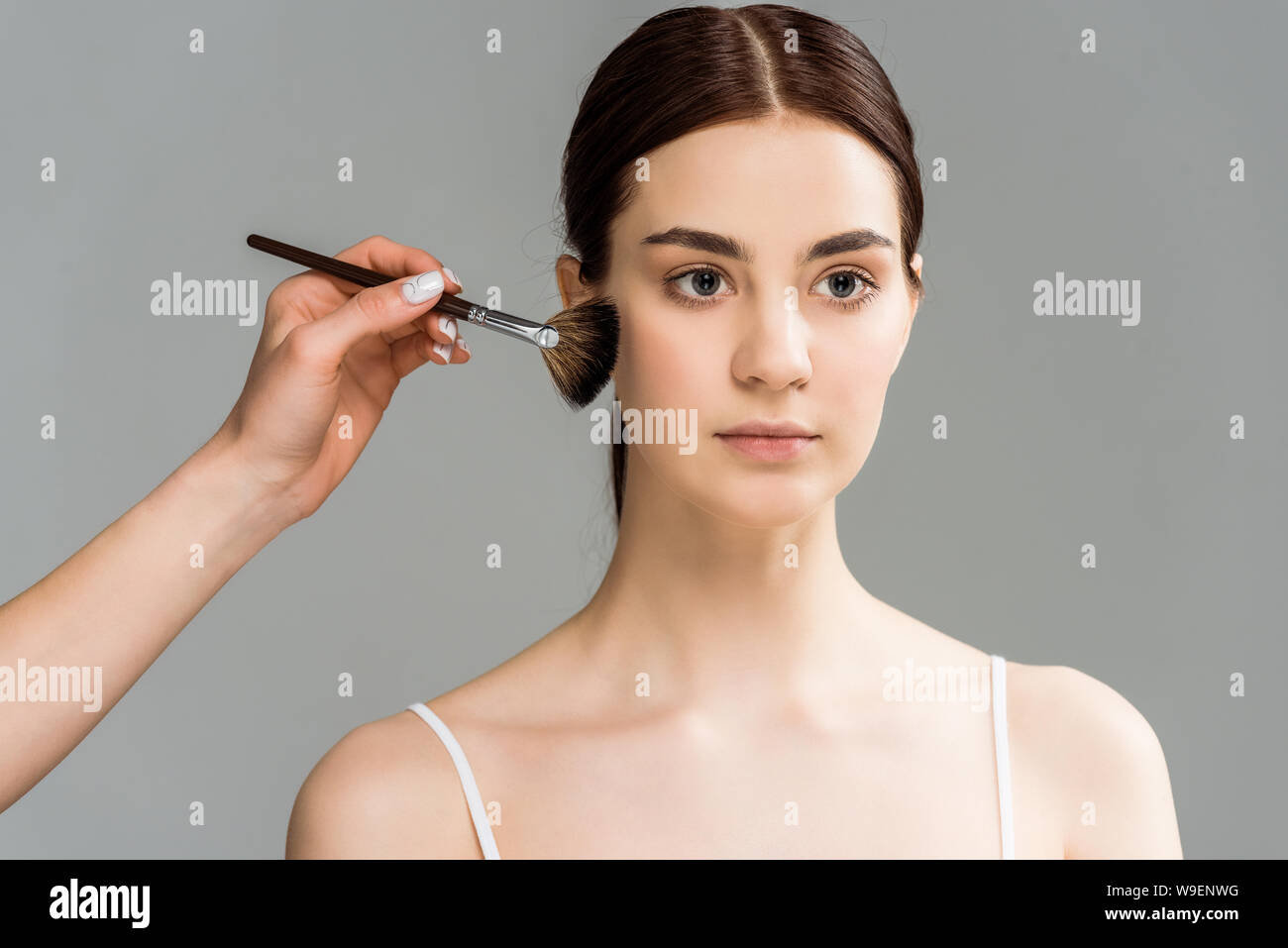 Vista recortada de maquillaje artista sosteniendo el cepillo cosmético cerca de la mejilla de la mujer hermosa aislado en gris Foto de stock