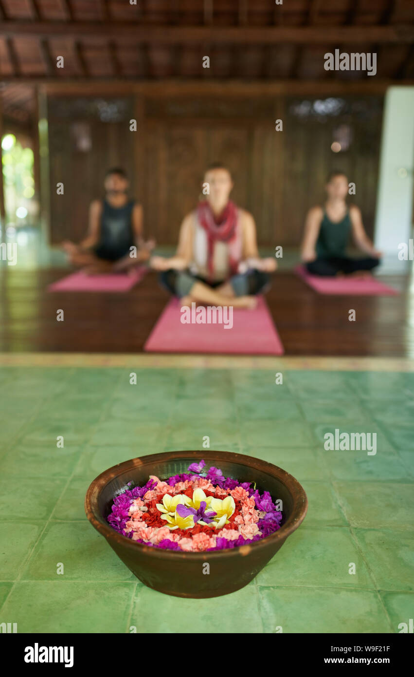 Cierre de la ofrenda de flores a la oración en el templo tradicional indonesio de Bali con tres diferentes personas meditando en segundo plano. Foto de stock