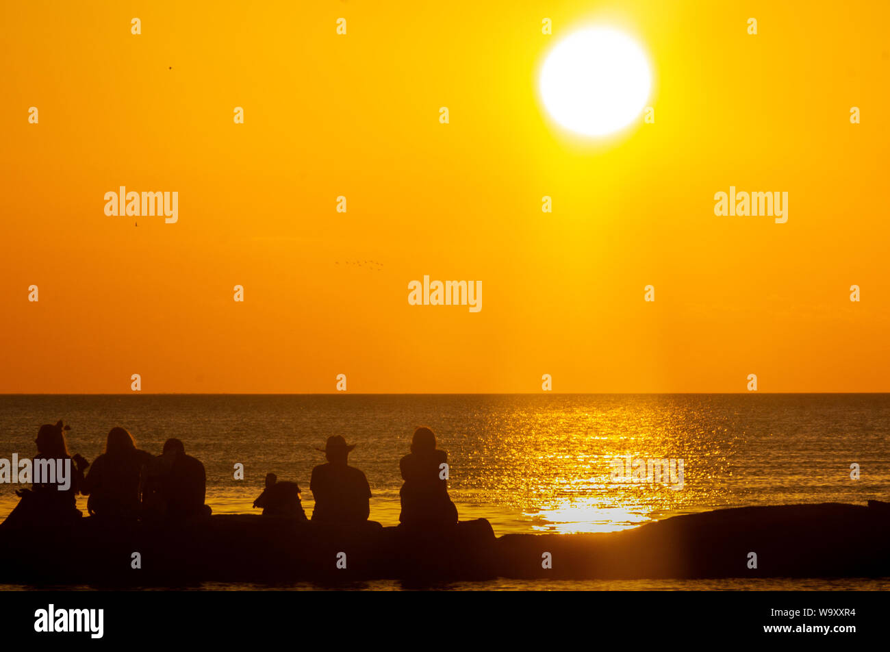 Silueta personas sentadas sobre su espalda en un rompeolas viendo la puesta de sol Foto de stock