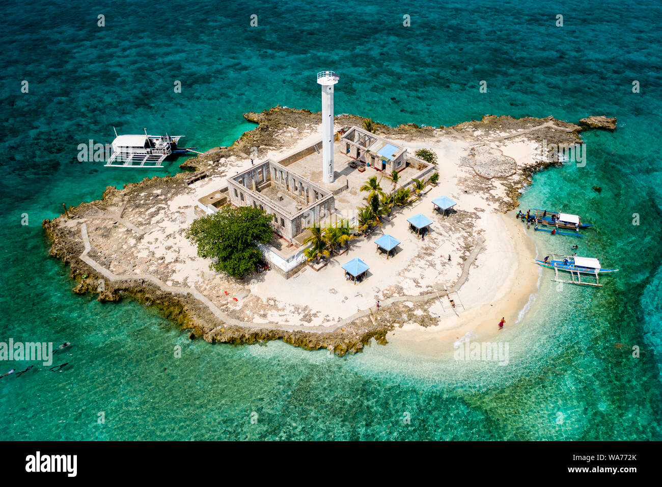 Drone vista aérea de embarcaciones tradicionales junto a una pequeña isla tropical con un faro, rodeada por arrecifes de coral (Isla Capitancillo, Filipinas) Foto de stock