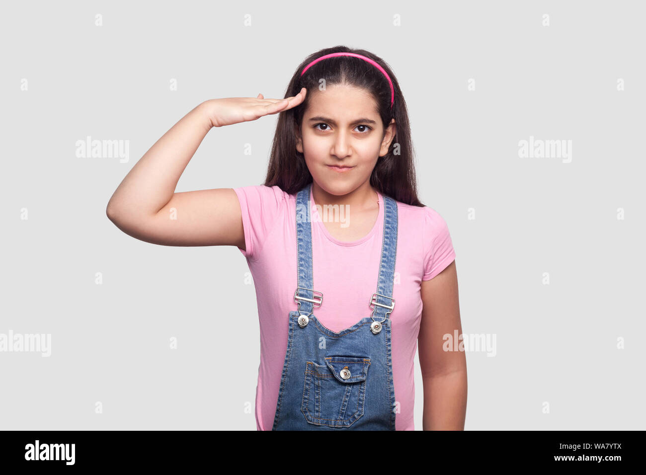 Sí señor. Retrato de grave joven morena en rosa t-shirt y un mono azul de pie con saludar el gesto y mirando a la cámara indoo atento. Foto de stock