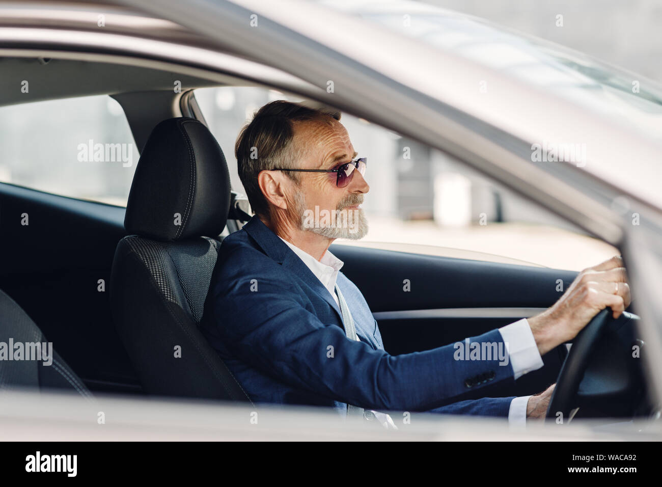 Empresario Senior conduciendo un coche. Vista lateral de un hombre en traje formal sentados en el vehículo. Foto de stock