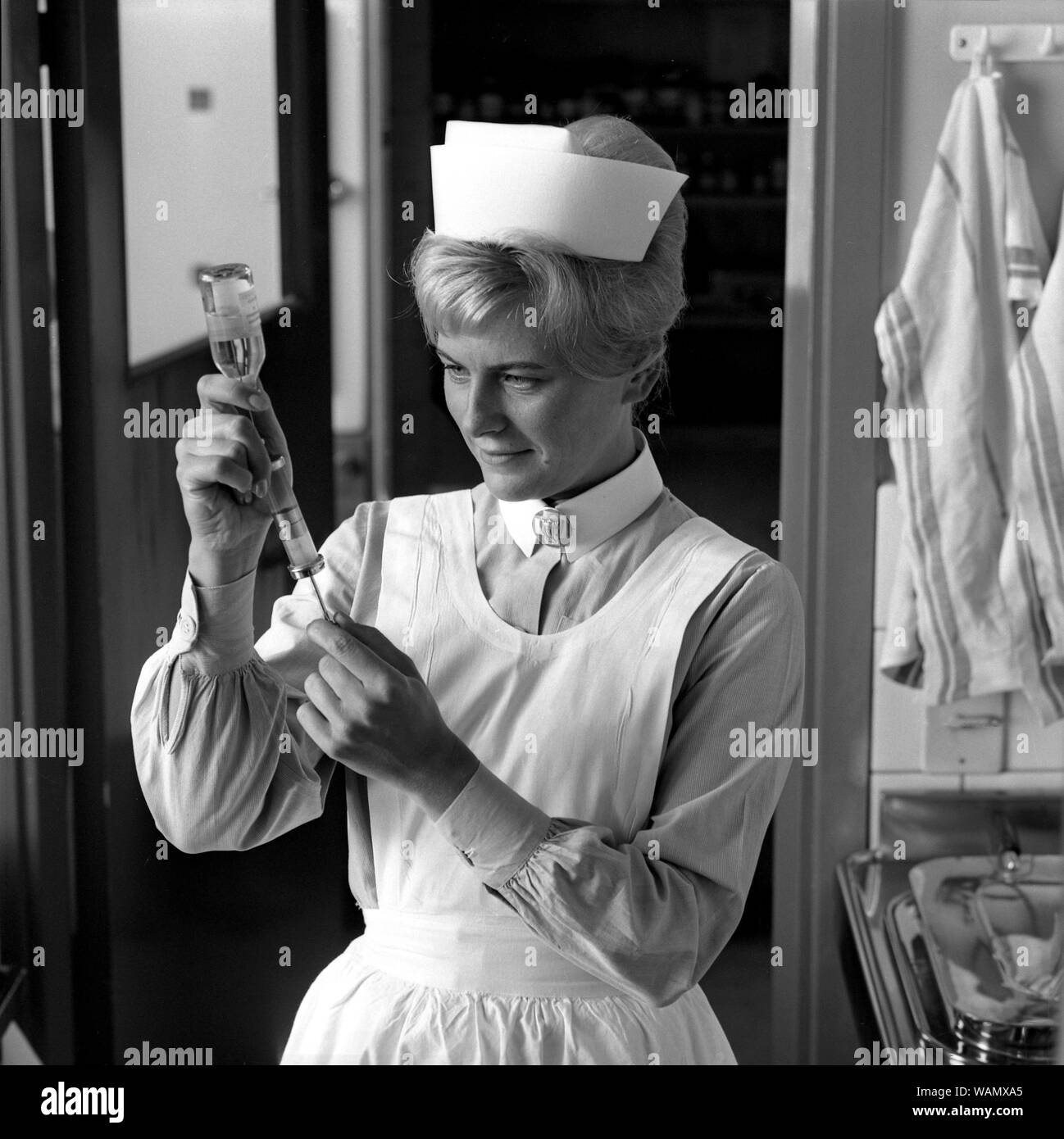 Enfermera en el decenio de 1960. Una mujer en un uniforme de enfermera está preparando una inyección y aspira el líquido de una botella de medicamento en una jeringa. septiembre de 1963. Foto de stock