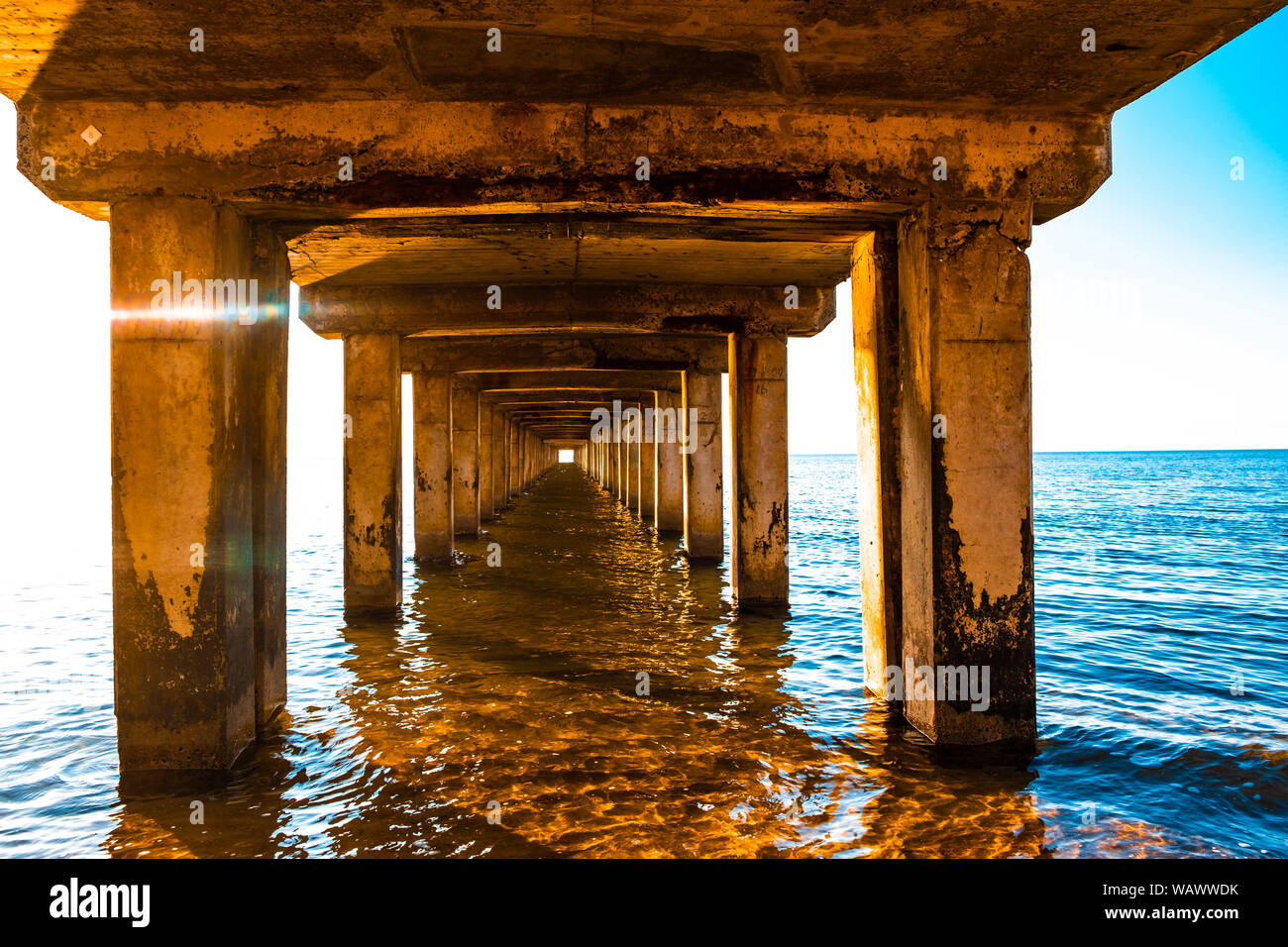 Perspectiva decreciente de pilares de madera debajo del viejo muelle largo de pie en el agua del océano al atardecer Foto de stock