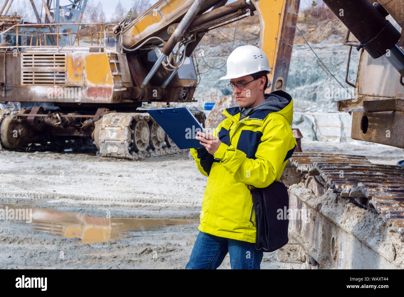 Macho geólogo o ingeniero de minas escribe algo en un mapa-caso en medio de una cantera con los equipos de construcción Foto de stock