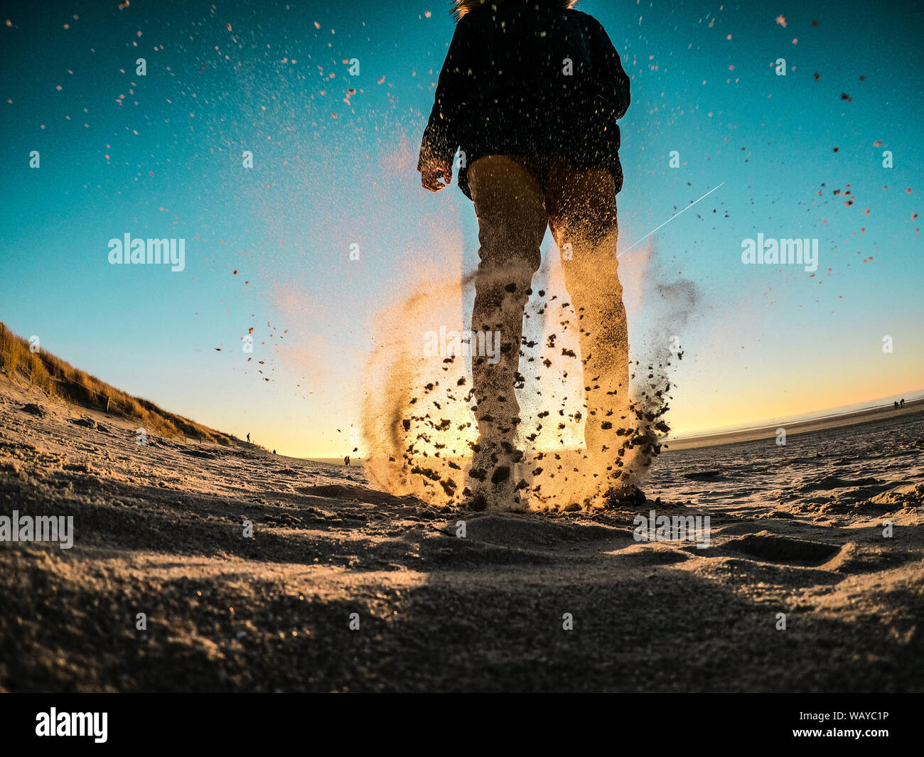 Chaqueta de invierno hombre vestido con pantalones de color naranja y golpeando la arena de la playa al atardecer Foto de stock