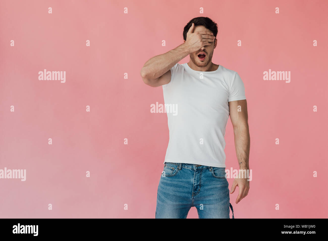 Vista frontal de conmocionado hombre muscular en la camiseta blanca que cubre los ojos con la mano aislados en rosa Foto de stock