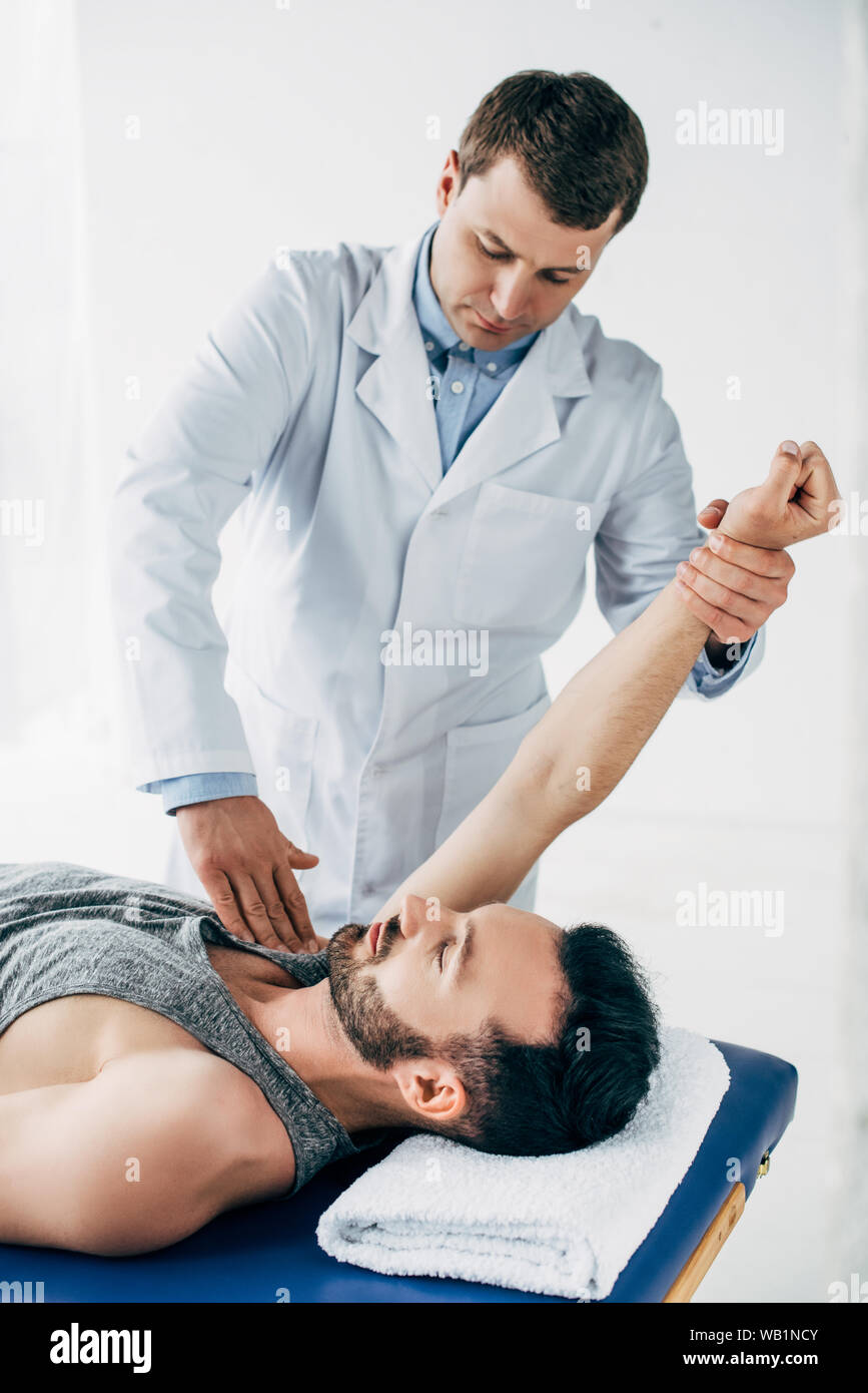 Quiropráctico estirando el brazo de guapo paciente tumbado en la mesa de masajes con toallas en el hospital Foto de stock