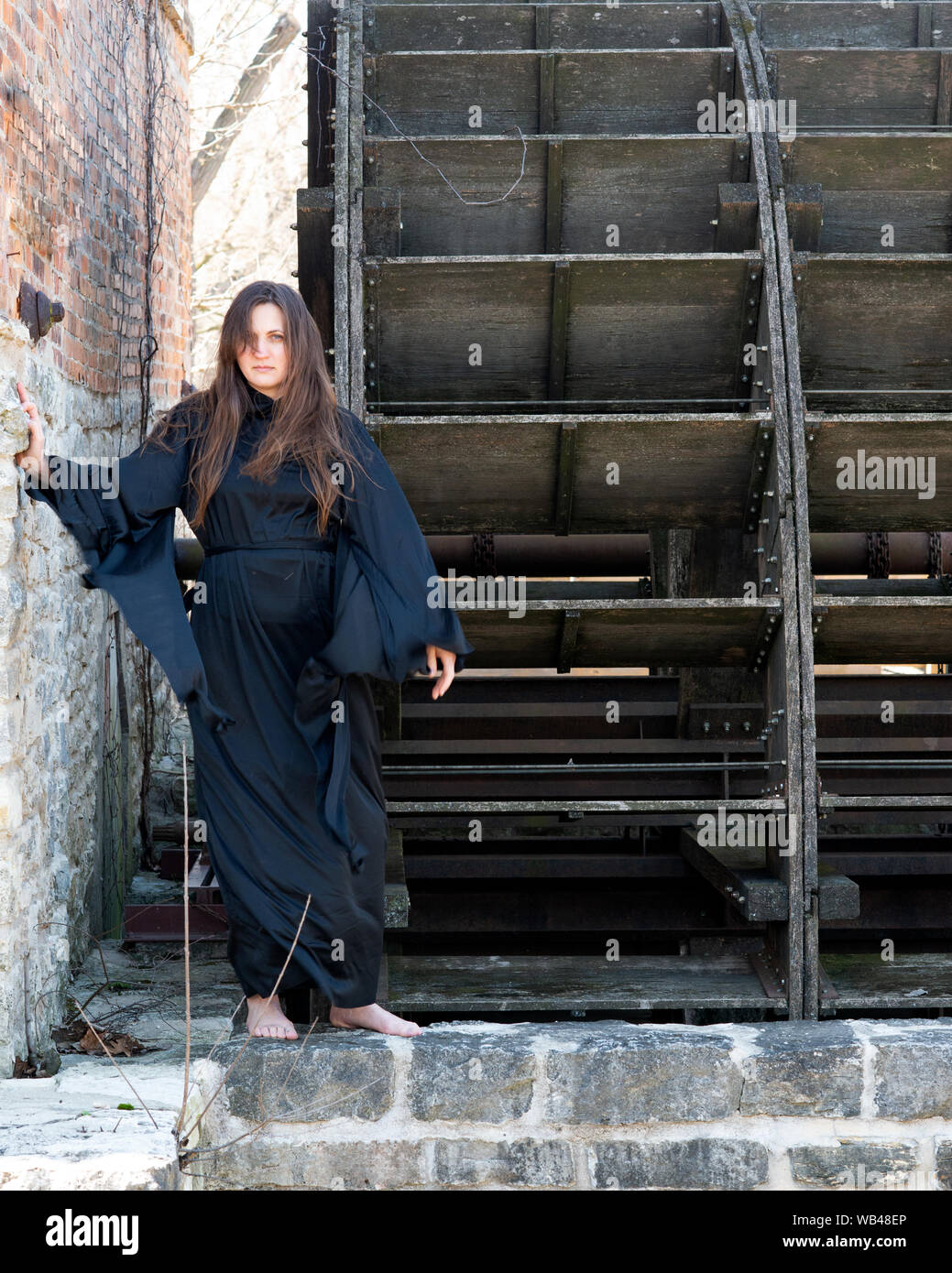 Mujer joven con largo cabello oscuro en túnicas negras en frente de un antiguo molino de agua de madera. Las Brujas. Concepto de Halloween. La brujería y la magia. Foto de stock
