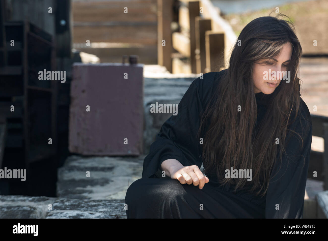 Mujer joven con largo cabello oscuro en túnicas negras en frente de un antiguo molino de agua de madera. Las Brujas. Concepto de Halloween. La brujería y la magia. Foto de stock