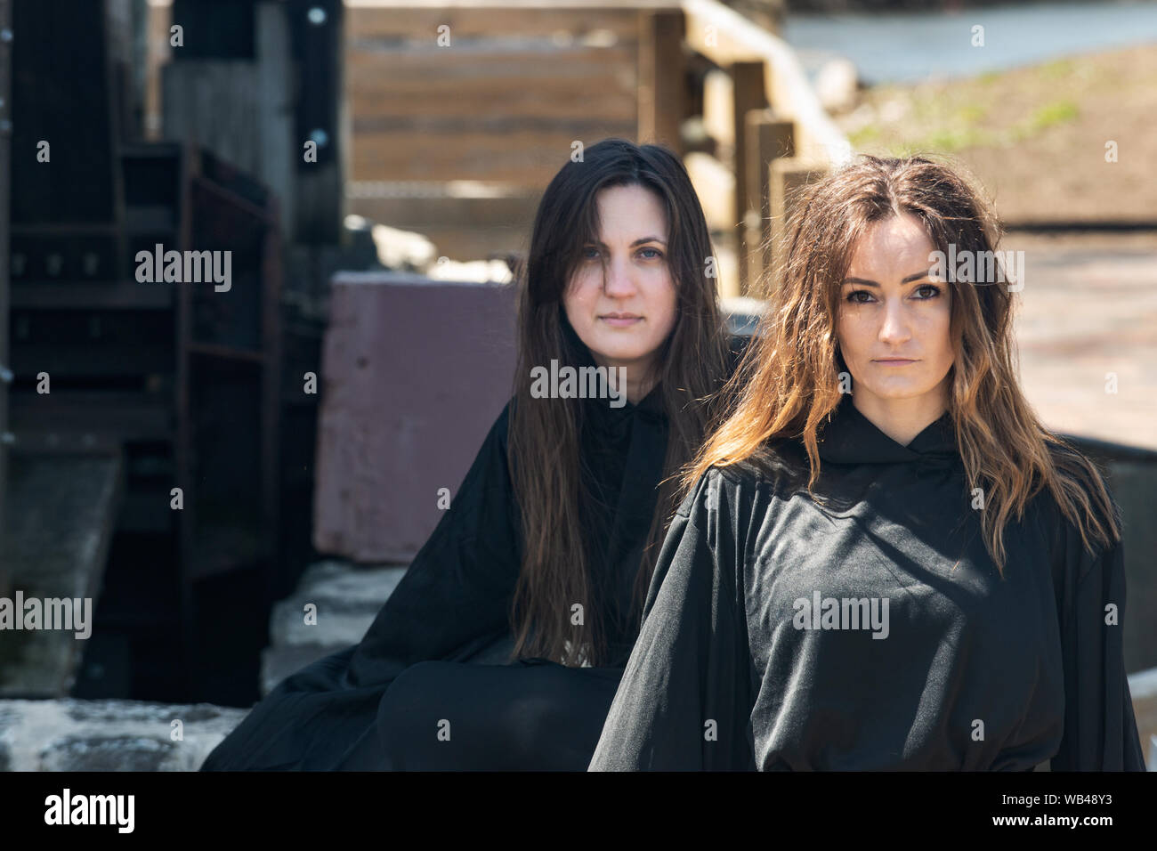 Las mujeres jóvenes con pelo largo oscuro en túnicas negras en frente de un antiguo molino de agua de madera. Las Brujas. Concepto de Halloween. La brujería y la magia. Foto de stock