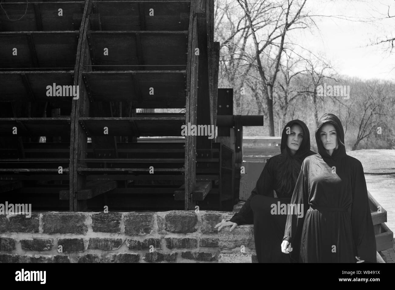 Las mujeres jóvenes con pelo largo oscuro en túnicas negras en frente de un antiguo molino de agua de madera. Las Brujas. Concepto de Halloween. La brujería y la magia. Foto de stock