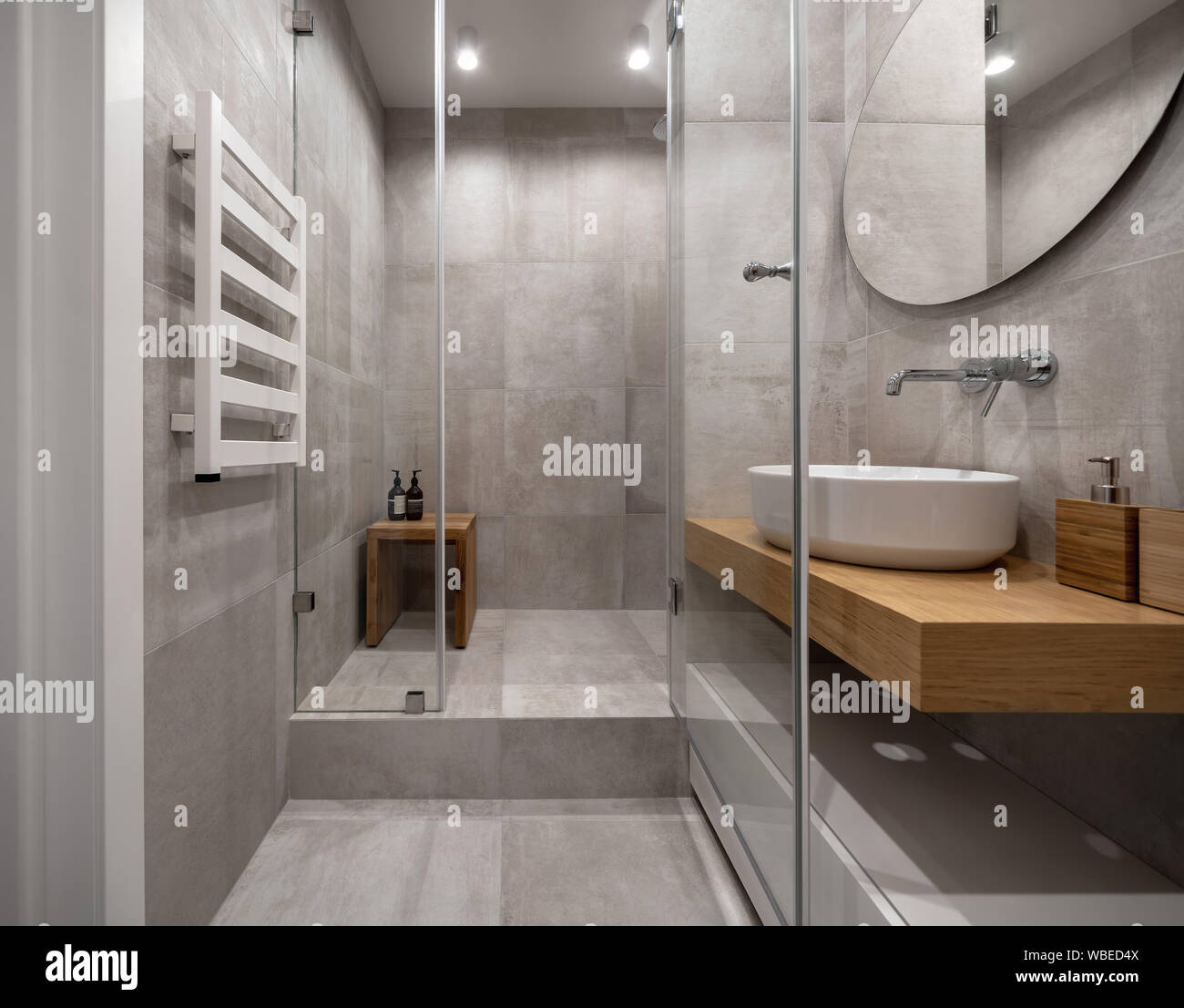 Cuarto de baño en un estilo moderno con paredes de baldosas color beige  texturado y luminosas lámparas. Hay una cabina de ducha de cristal, estante  de madera con un lavabo blanco y