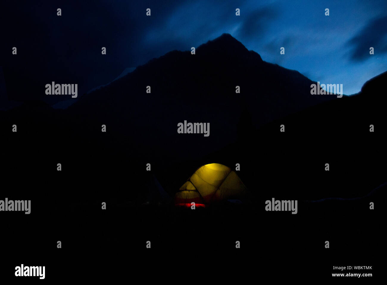 Vista nocturna de camping: una carpa iluminada desde el interior, sihoutte de montañas en el fondo Foto de stock