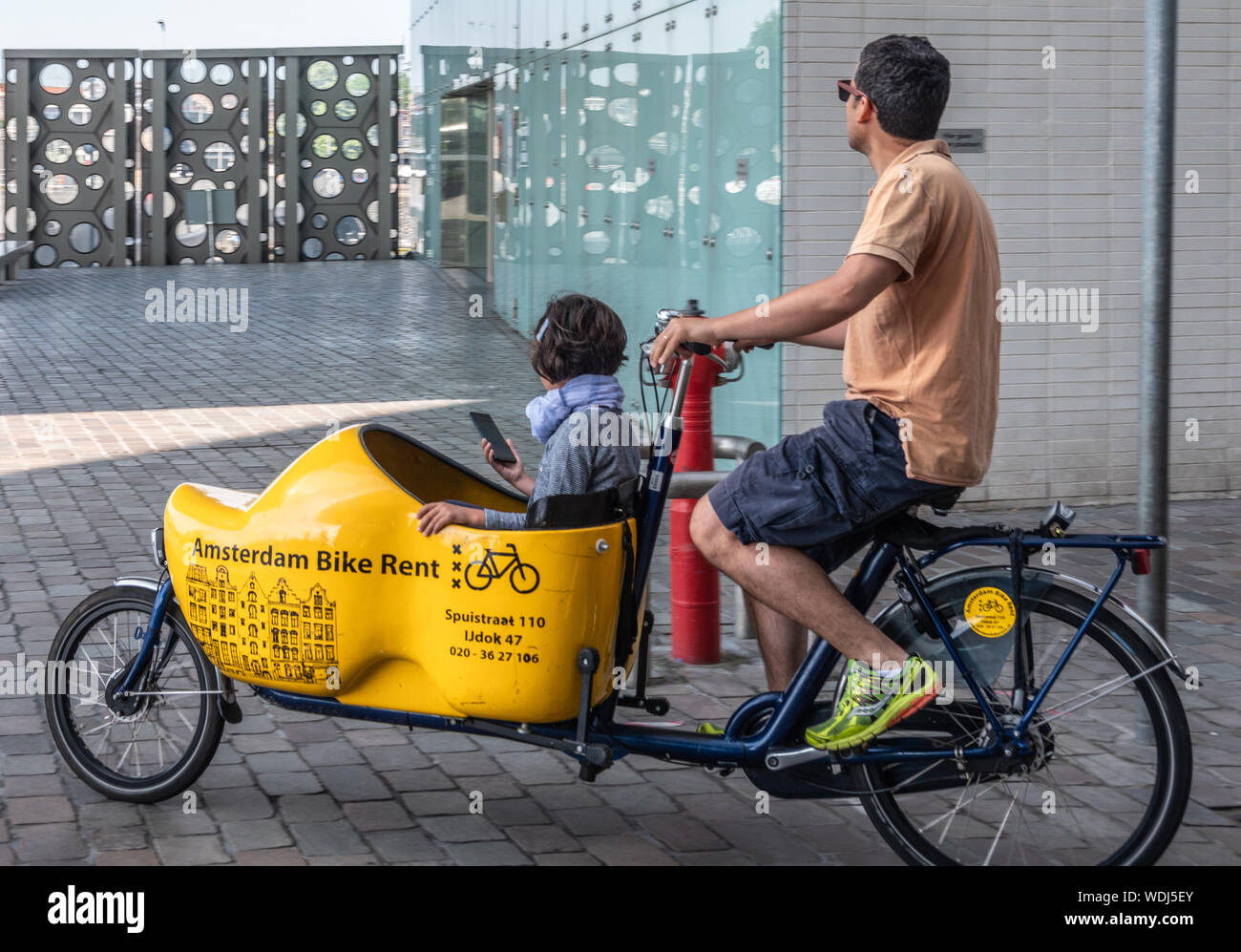 Amsterdam, Holanda - 30 de junio de 2019: Primer plano de bakfiets,  delantero-trunk bicicleta para transportar una carga, un niño aquí. El  hombre tal como se ve en IJdok pedaleando. Tr Fotografía