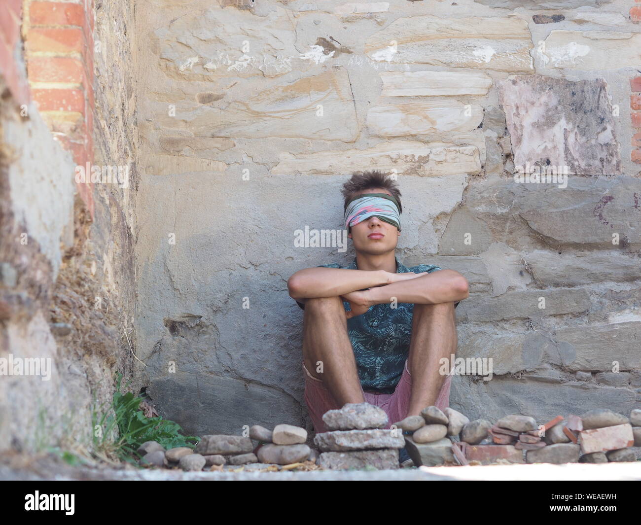 Hombre con los ojos cubiertos sentado contra la vieja pared desgastada Foto de stock