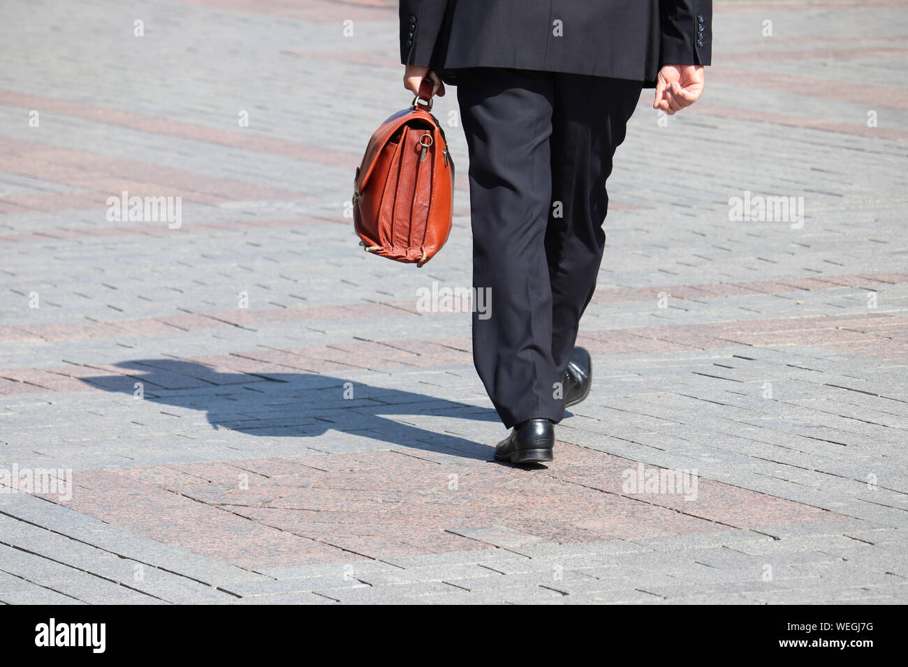Hombre en un traje de negocios llevando maletín de cuero caminando por una calle, sombra negra sobre el pavimento. Concepto de empresario, funcionario, político, carrera Foto de stock