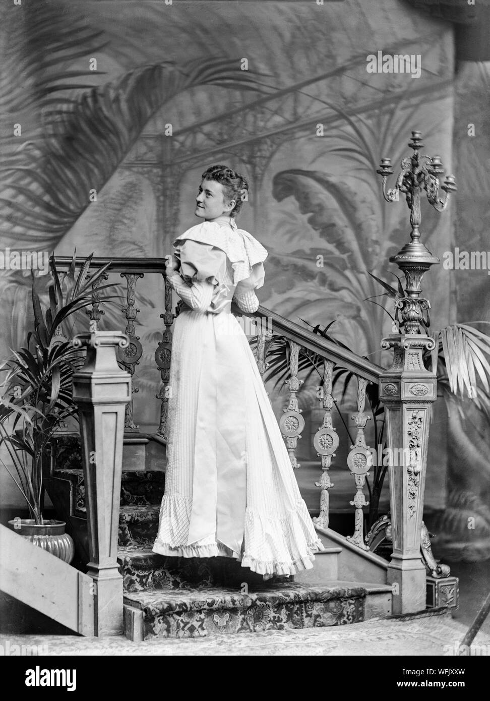 Un vintage victoriano tardío o principios Edwardian fotografía en blanco y negro mostrando una señorita en un fino vestido a la moda de la época, posando en un estudio fotográfico, con su espalda ligeramente hacia la cámara, como ella mira sobre su hombro. Ella está de pie sobre un conjunto de escaleras. Foto de stock