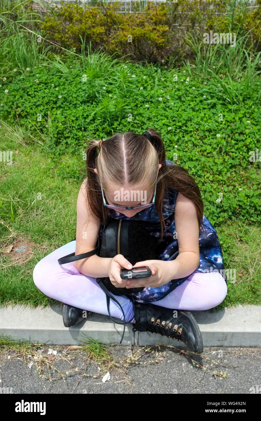 Un joven pre-adolescentes caucásicos niña jugando con su teléfono móvil mientras está sentado en el bordillo de la acera. Foto de stock