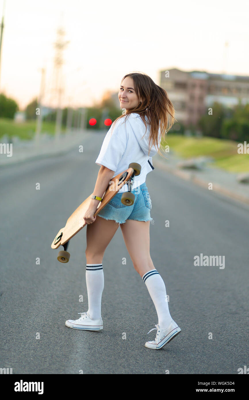 Retrato de una niña de la calle en ropa casual y longboard en su mano está  en el patio de recreo, mira a la cámara y poses. Niña modelo con ride bo
