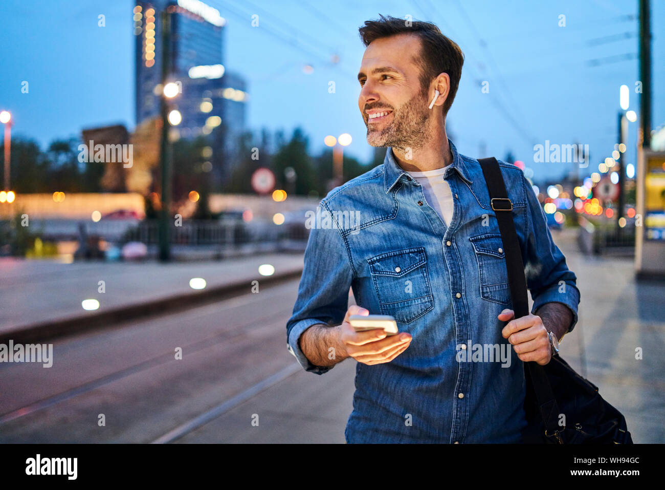 Hombre sonriendo con smartphone y escuchar música a través de unos auriculares inalámbricos mientras esperan en la parada de tranvía de conmutar durante la noche después del trabajo Foto de stock