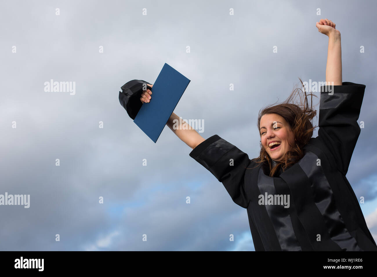 Bastante joven, celebrando con alegría su graduación - difusión amplia sus brazos, sosteniendo su diploma, saboreando su éxito (la imagen en tonos de color; shal Foto de stock