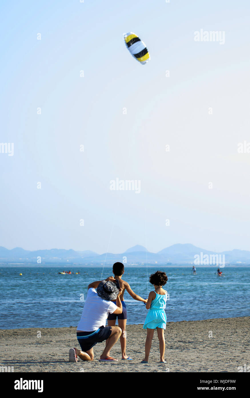 Familia Feliz en verano en la playa. Niños jugando con un cometa con su padre en la orilla del mar. La paternidad con su hijo e hija volar cometas, España, 2019. Foto de stock