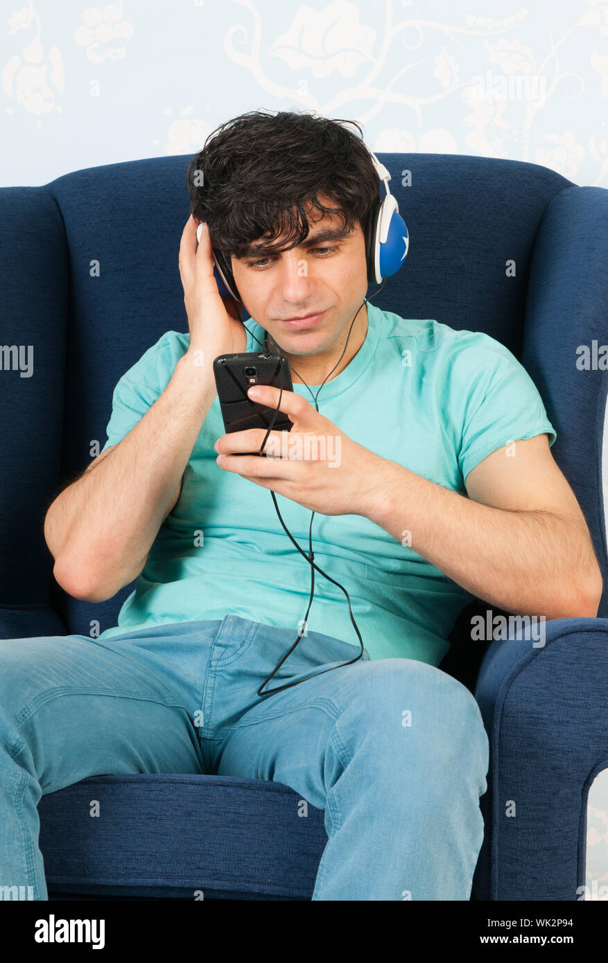 Joven sentado en un sillón azul y escuchando música con el teléfono inteligente. Foto de stock