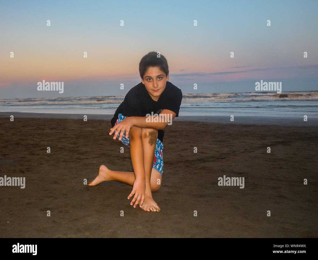 Retrato de niño arrodillado en la playa contra el cielo durante la puesta de sol Foto de stock