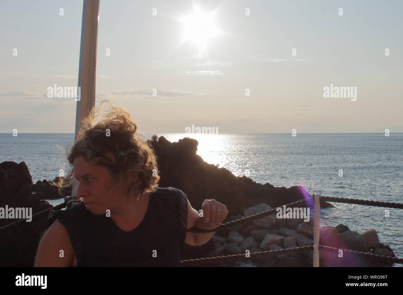 Una mujer rubia caucasic viendo un velero en el horizonte durante la puesta de sol en el bar de la playa Foto de stock