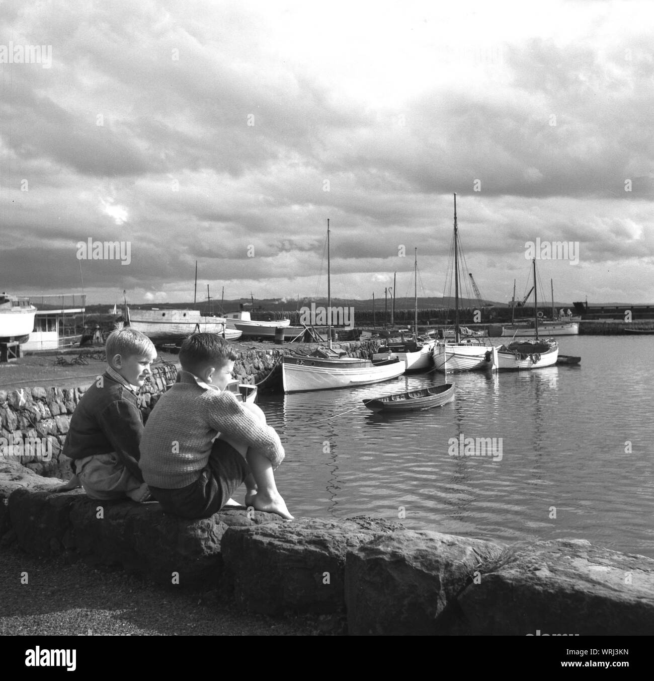 1950s, históricos, dos muchachos sentados en el muro del puerto mirando los barcos y el mar, Co. de Antrim, Irlanda del Norte. Foto de stock