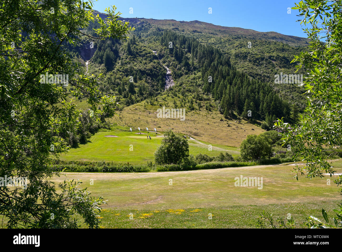 Vista panorámica de un paisaje de montaña con un campo de golf rodeado por bosques de pinos y cascadas en verano, Val Ferret,Courmayeur (Valle de Aosta, Italia Foto de stock