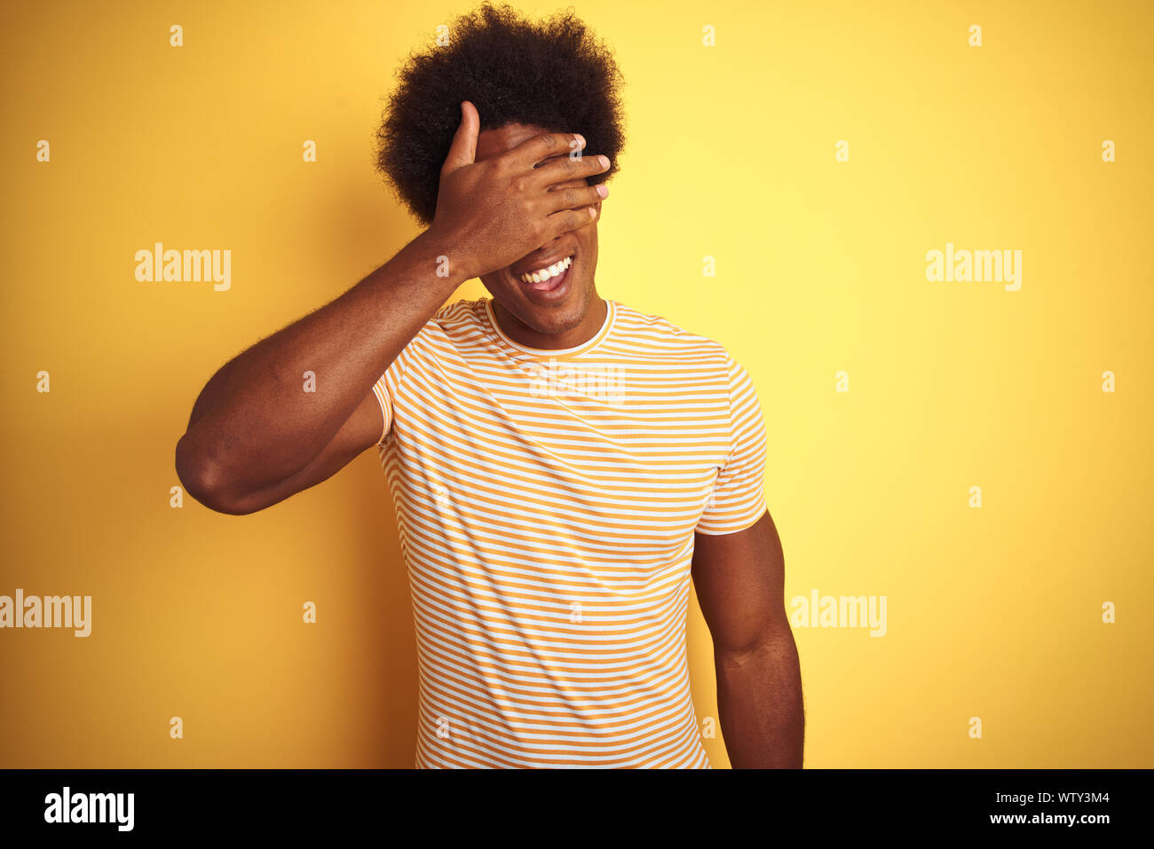 Hombre americano con el cabello afro vistiendo camiseta de rayas parado  sobre fondo amarillo aislado sonriendo y riendo con la mano sobre la cara  para tapar los ojos Fotografía de stock -