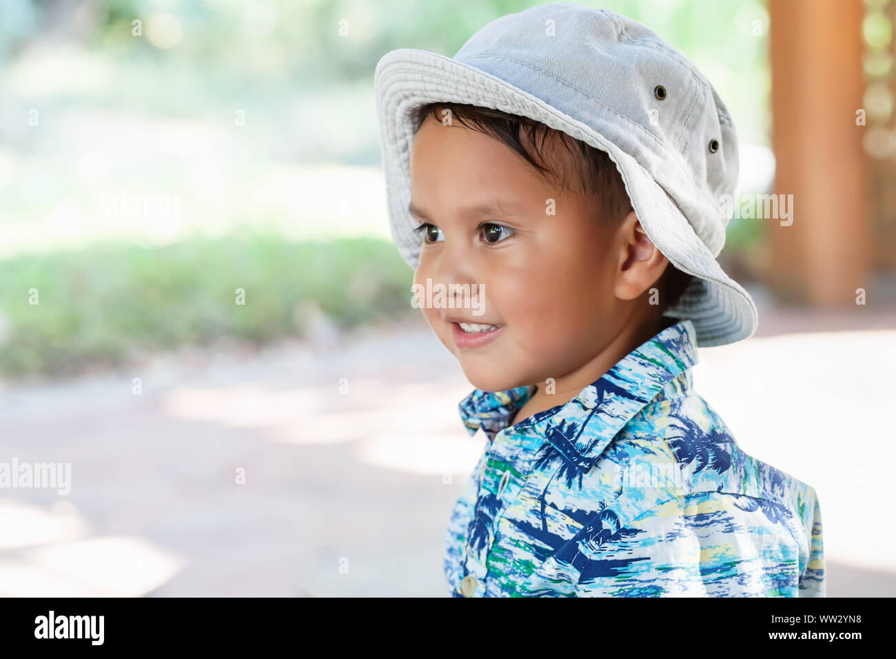 Un chiquillo; 3 año de edad, vistiendo un sombrero y una camiseta de impresión hawaiano, mirando en la distancia con una linda sonrisa en la cara. Foto de stock