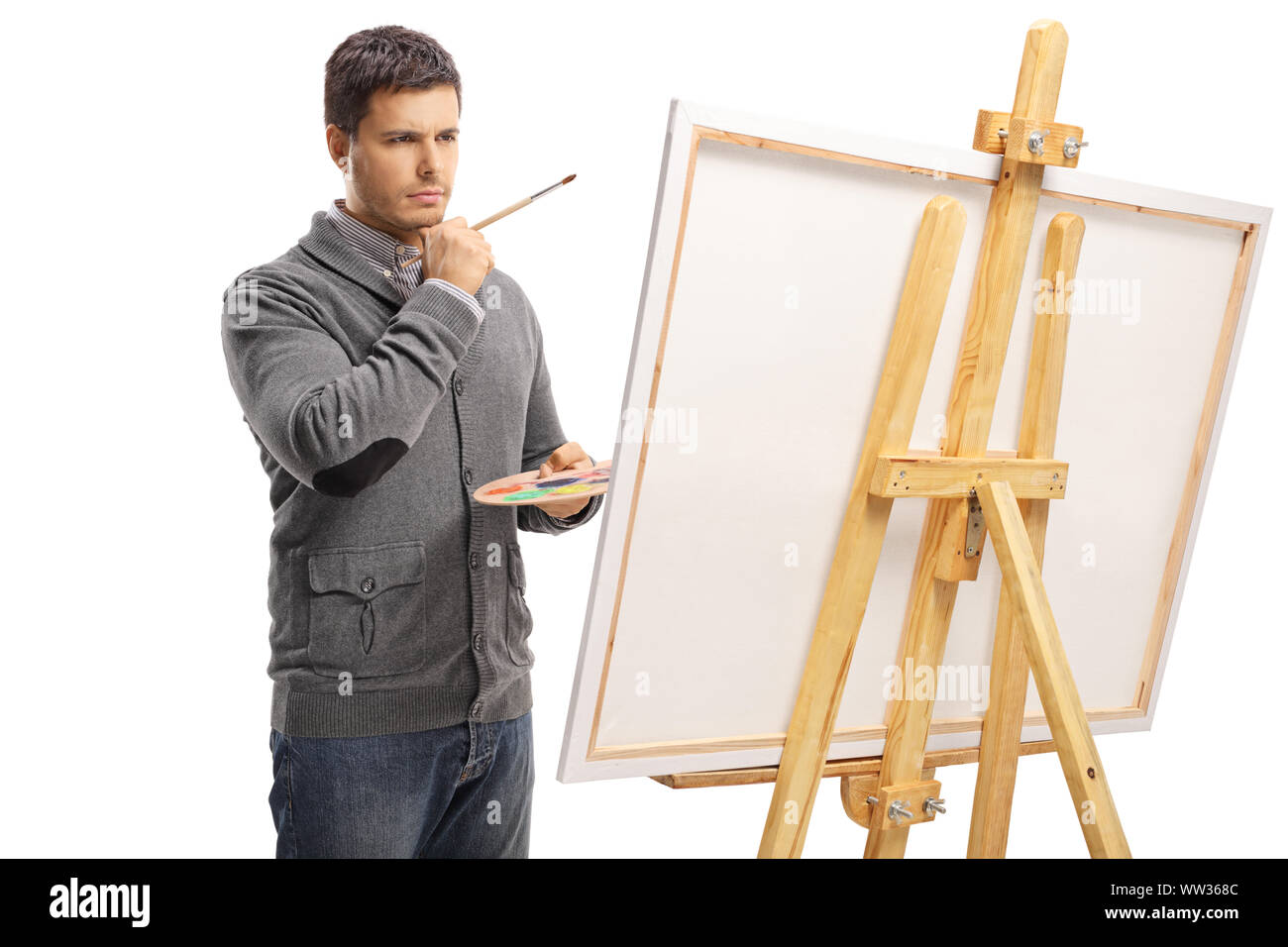 Pensativo hombre de pie delante de un lienzo de pintura con un pincel y paleta aislado sobre fondo blanco. Foto de stock