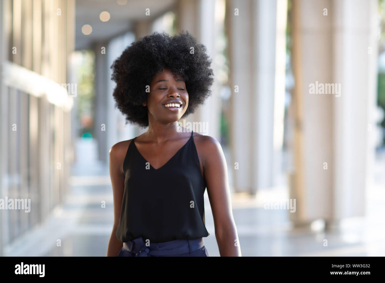 Sonriente joven mujer negra caminando por la ciudad y disfrutar los días soleados con los ojos cerrados Foto de stock