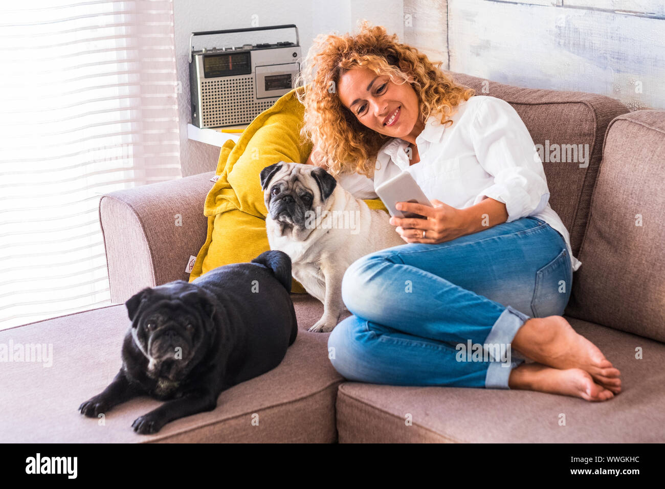 Mujer alegre en casa sentarse en el sofá con sus dos mejores amigos de pug perro cerca de ella - estilo de vida real de las personas con animales Foto de stock