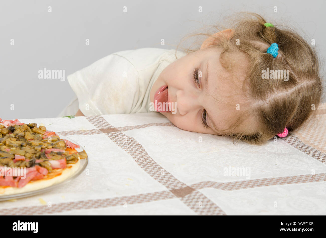Niña con lengua protuberante descansaba su cabeza sobre la mesa y mira la pizza Foto de stock