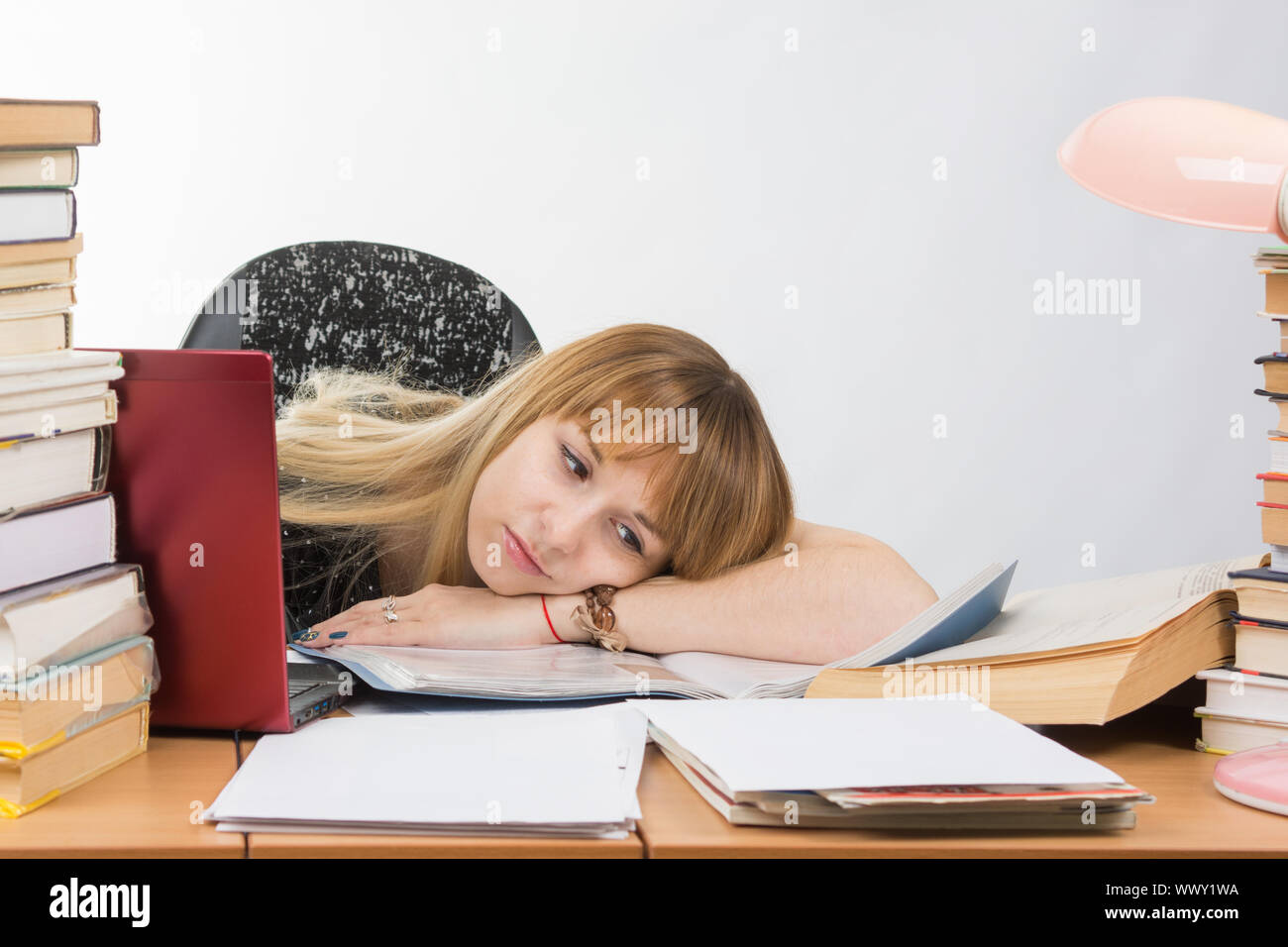 Chica estudiante puso su cabeza en sus manos repletas de libros y papeles tirados en el escritorio Foto de stock