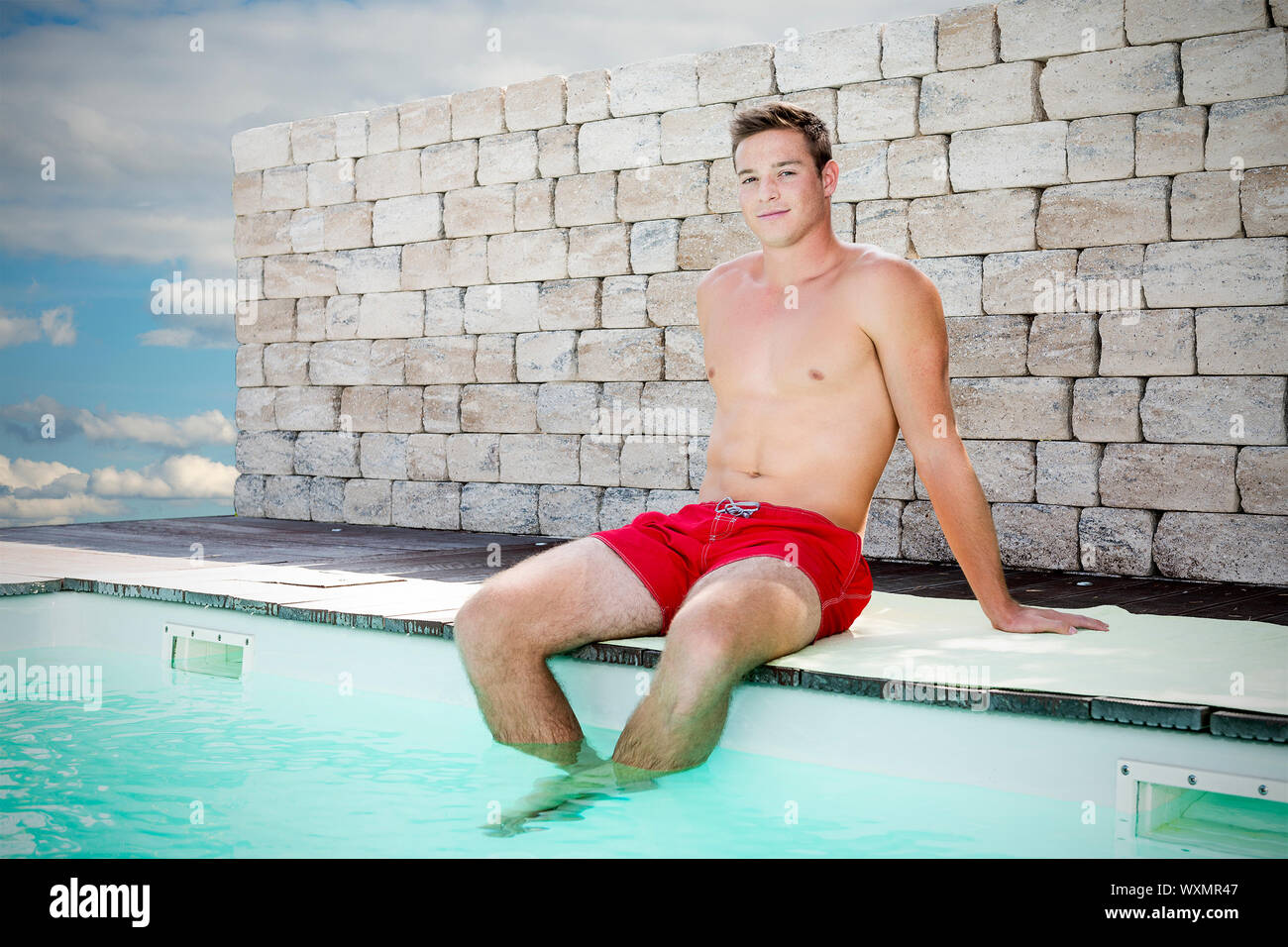 Joven apuesto hombre sentado en el borde de una piscina con shorts Foto de stock