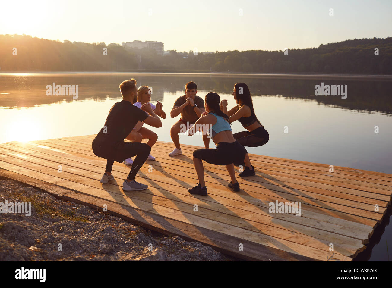 Un grupo de deportistas, hacer ejercicios en cuclillas en un parque junto al lago Foto de stock