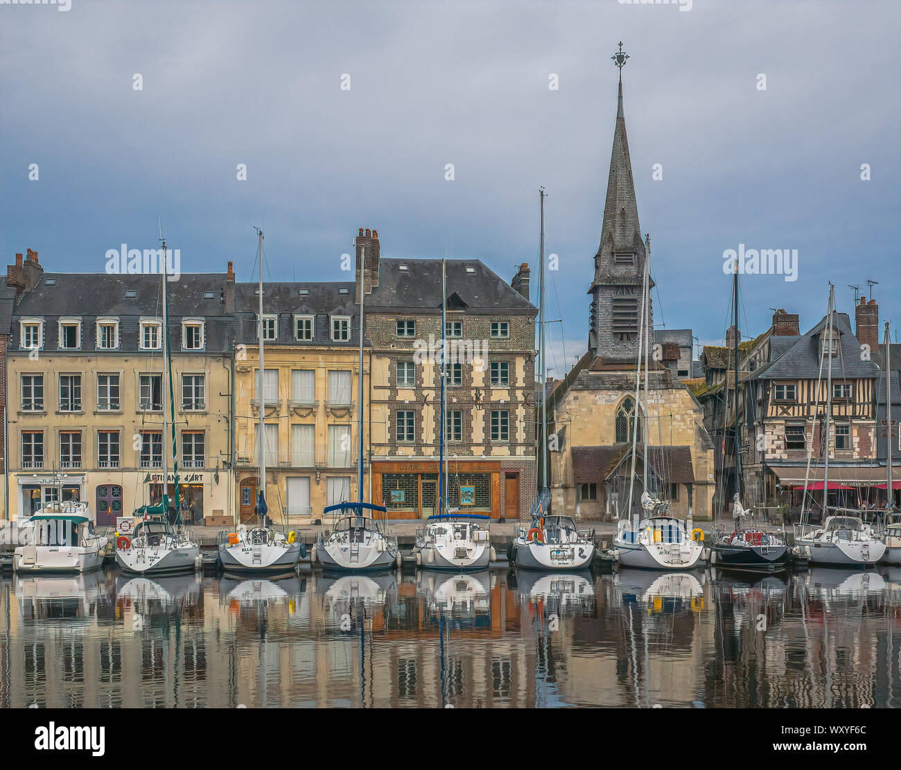 Línea de veleros el muelle debajo de los edificios históricos y la iglesia de Saint Etienne se reflejan en las quietas aguas del viejo puerto de Honfleur, Francia Foto de stock