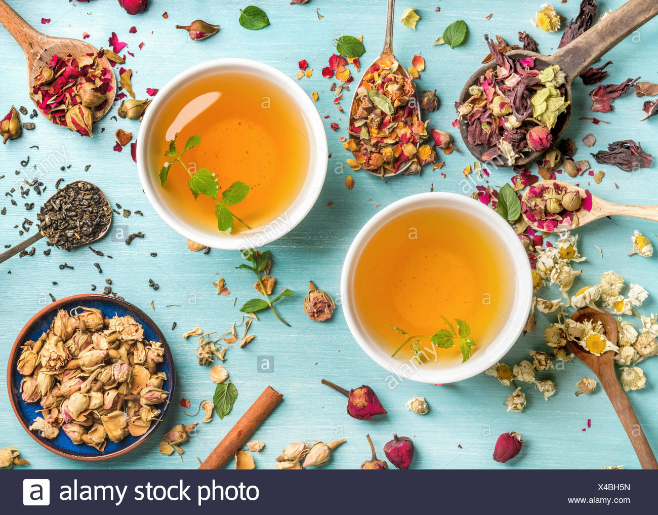 Dos tazas de té de hierbas saludables con menta, canela, rosa y flores secas de manzanilla en cucharas sobre fondo azul, vista superior Foto de stock