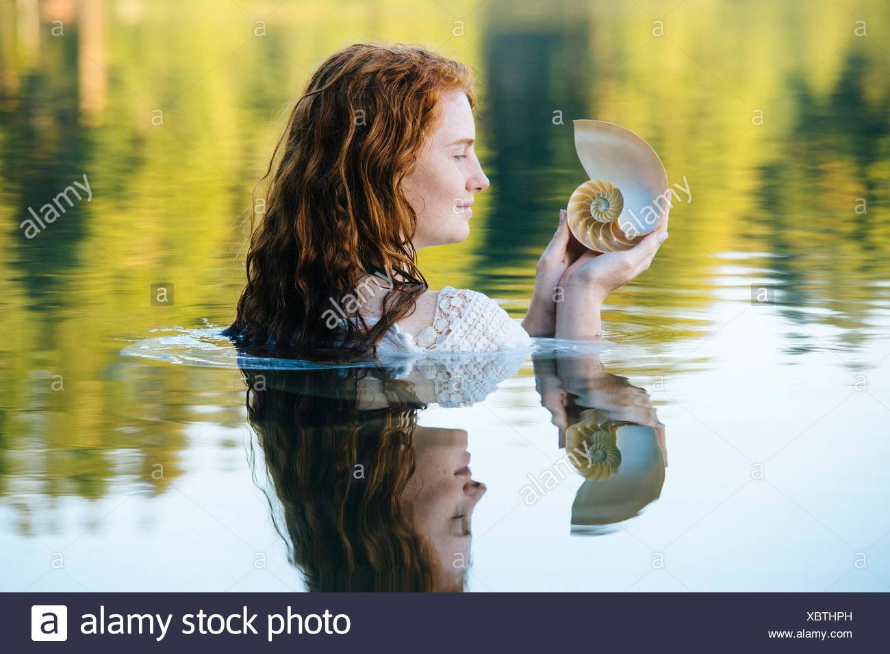 Cabeza y hombros de la mujer joven con largo pelo rojo en el lago contemplando el Seashell Foto de stock