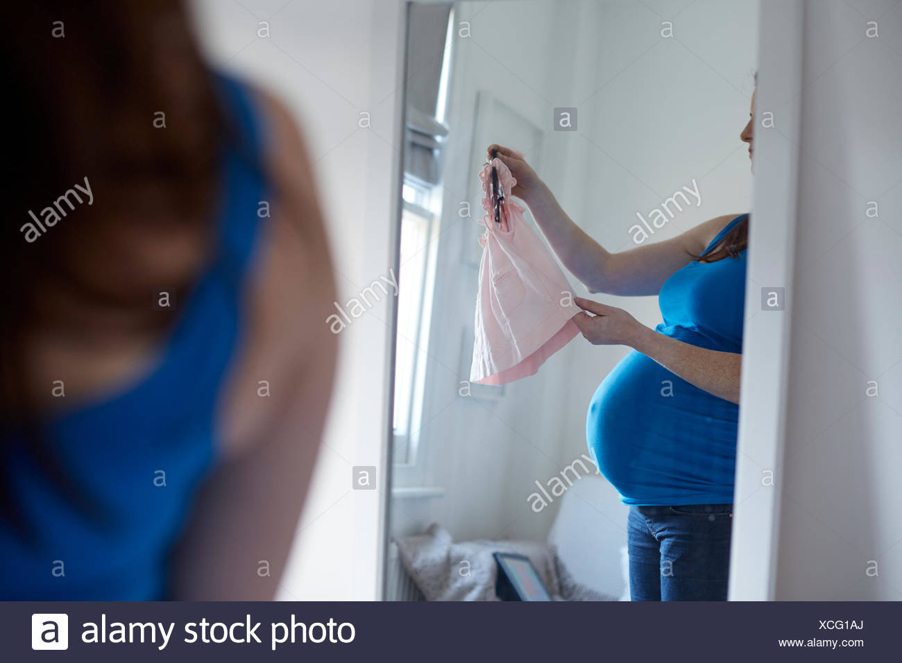 Imagen de espejo de la madre embarazada la celebración de ropa de bebé en la percha Foto de stock