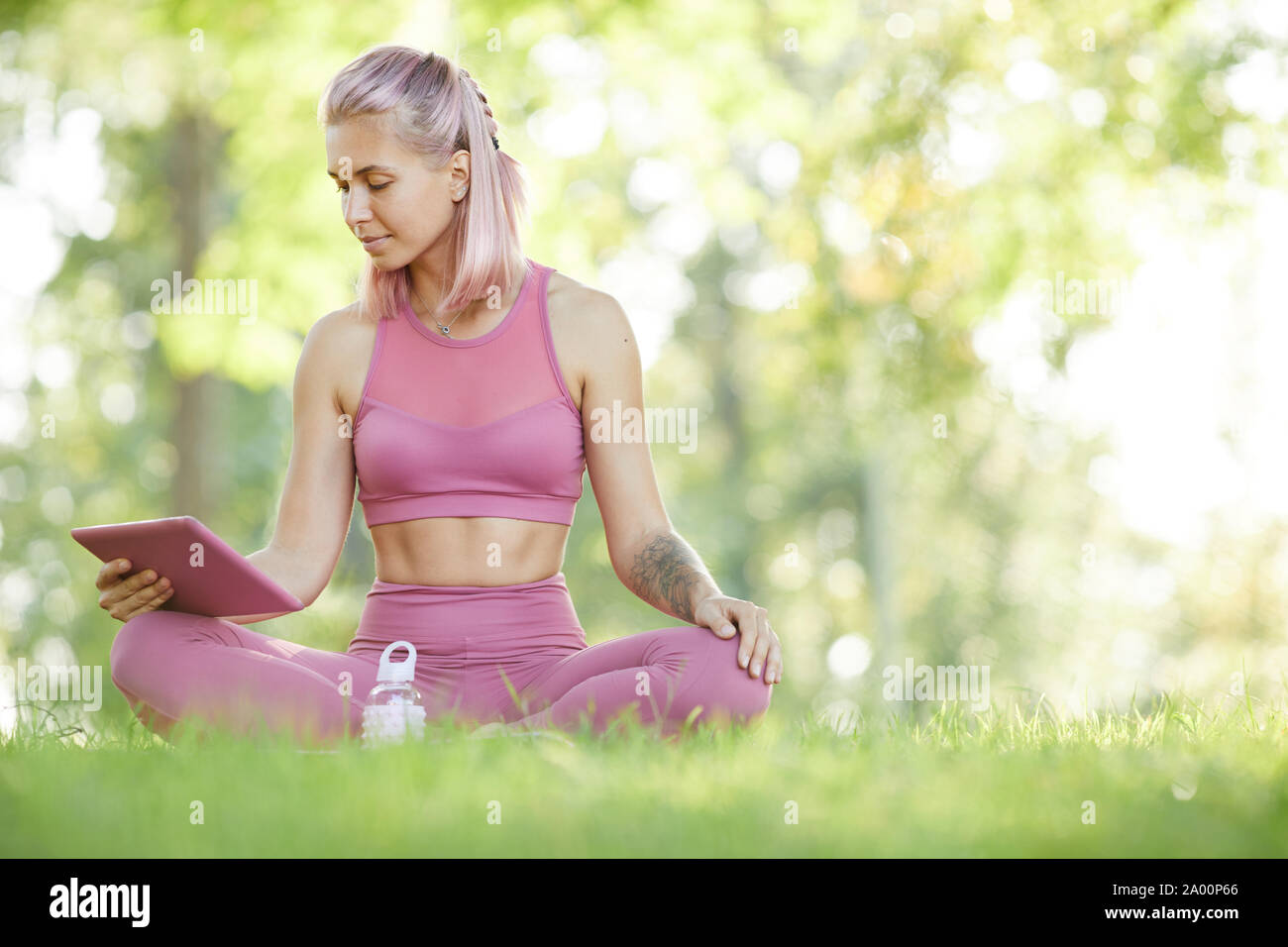 Young woman sitting on the grass and using digital tablet elle regardant des leçons en ligne des exercices sportifs en plein air Banque D'Images