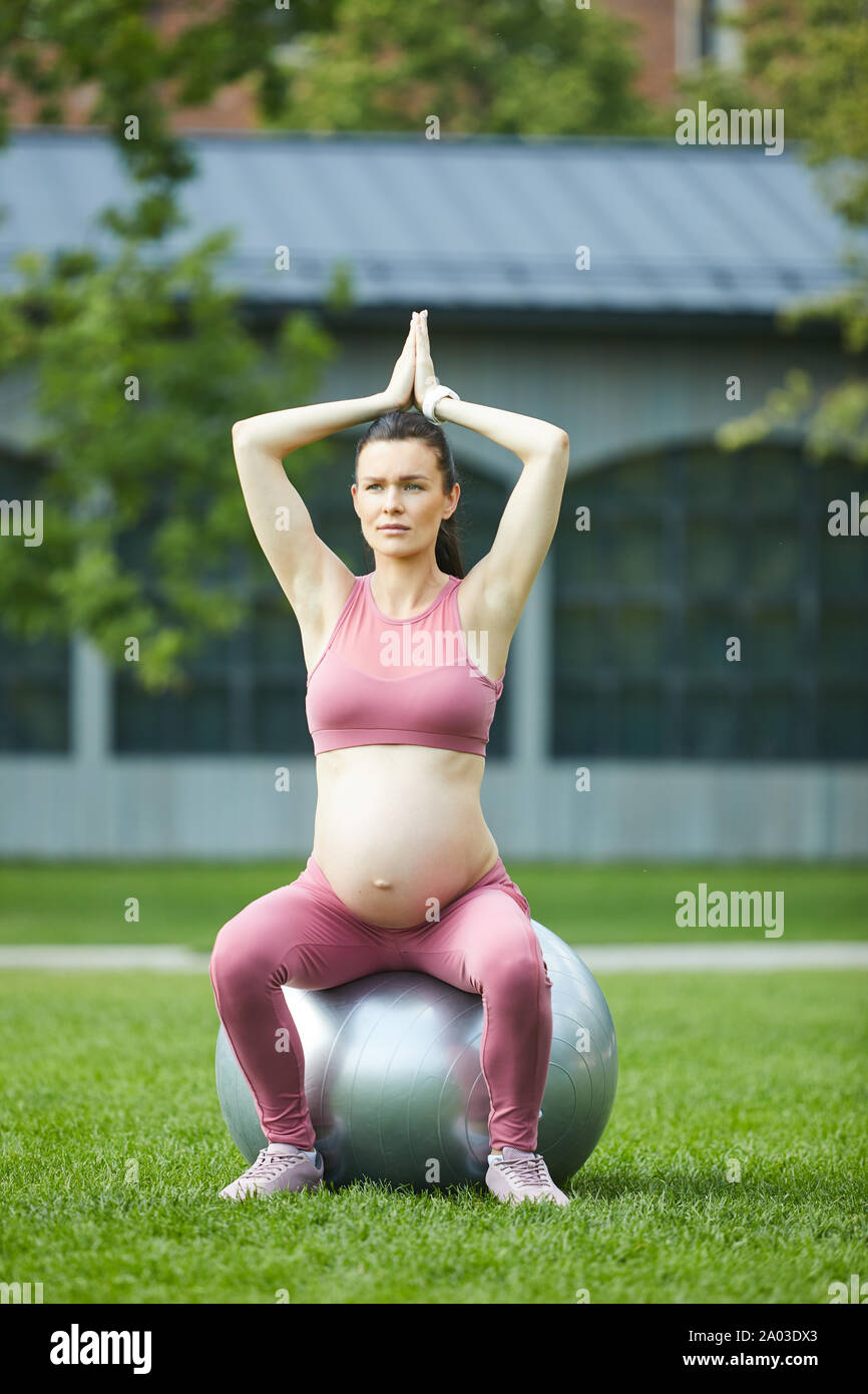 Young pregnant woman sitting on fitness ball a soulevé ses bras et faisant de l'exercice à l'extérieur dans le parc Banque D'Images